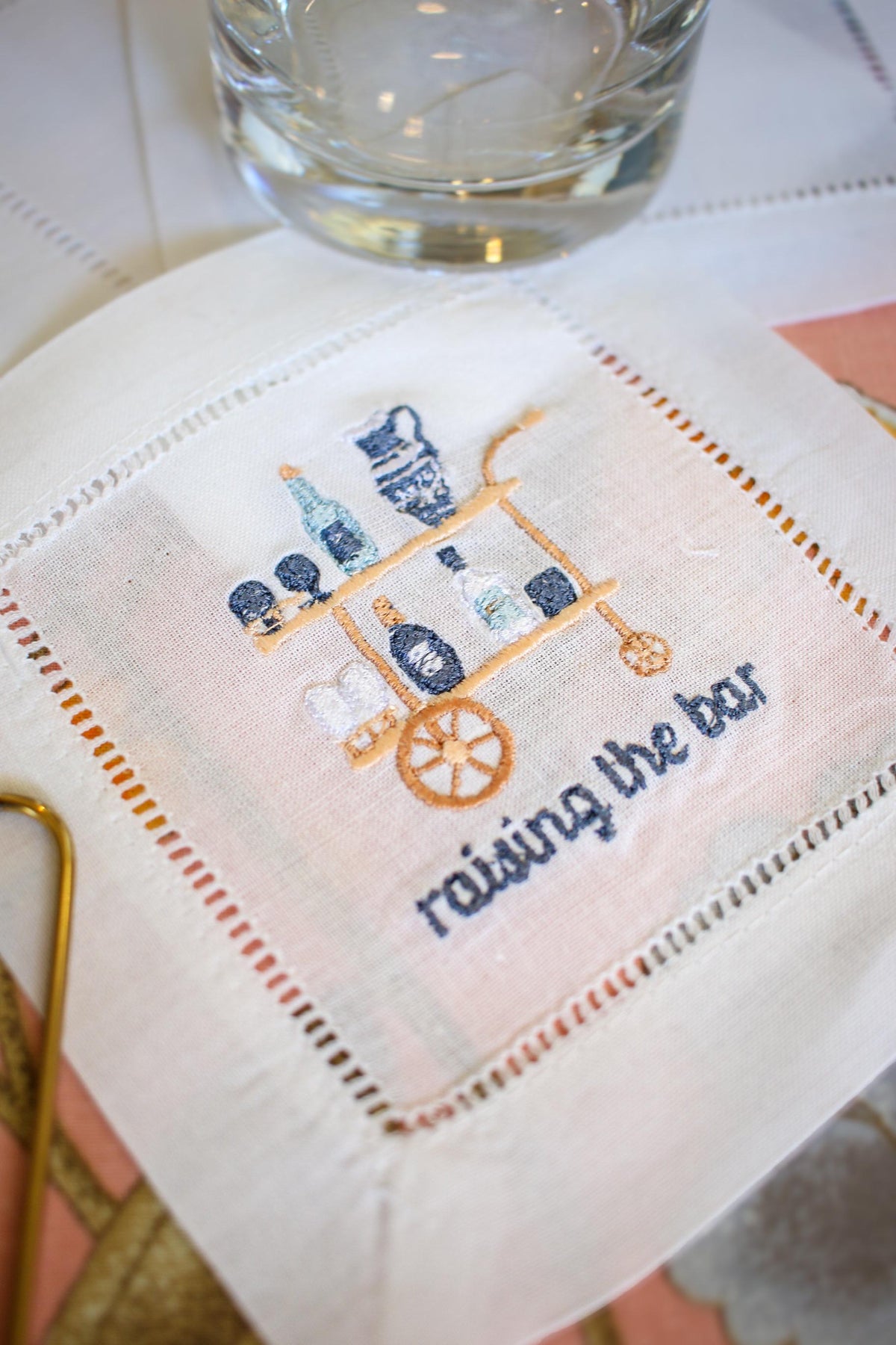 white hemstitch cocktail napkin with custom embroidery including a blue and gold bar cart and embroidered words below reading "raising the bar" in navy blue
