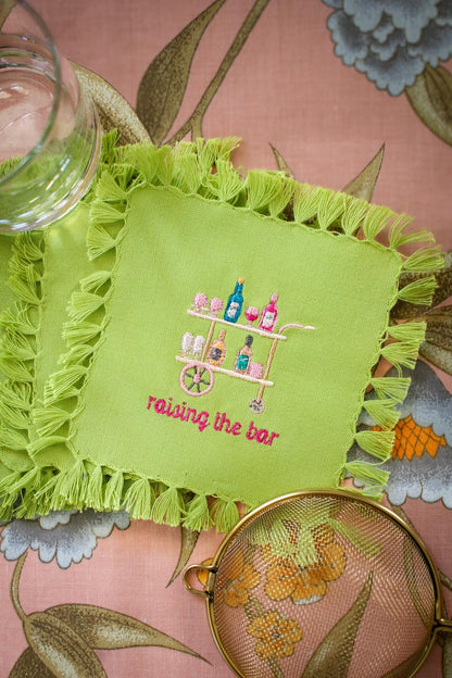 lime green cocktail napkin with tassel fringe and custom embroidery including a peach, pink, and blue bar cart and embroidered words below reading "raising the bar" in hot pink
