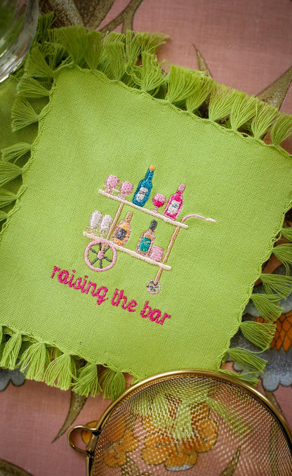 lime green cocktail napkin with tassel fringe and custom embroidery including a peach, pink, and blue bar cart and embroidered words below reading "raising the bar" in hot pink