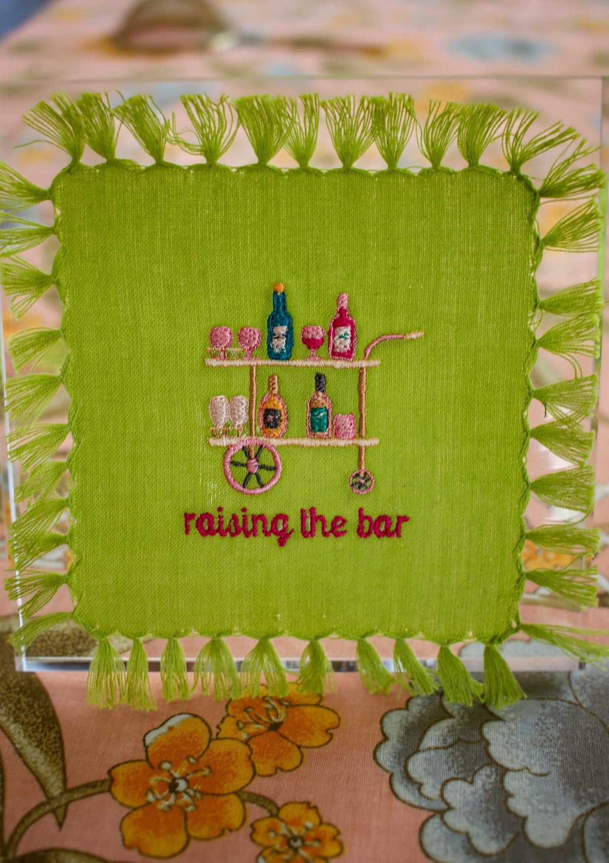 lime green cocktail napkin with tassel fringe and custom embroidery including a peach, pink, and blue bar cart and embroidered words below reading "raising the bar" in hot pink