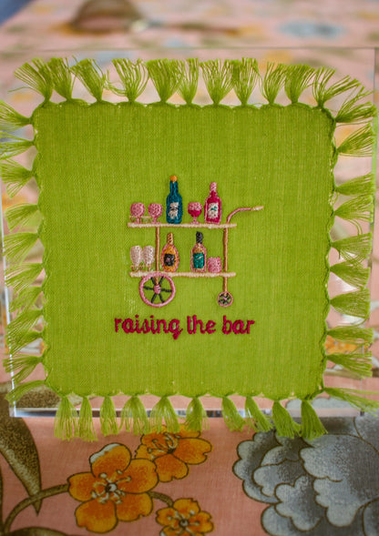 lime green cocktail napkin with tassel fringe and custom embroidery including a peach, pink, and blue bar cart and embroidered words below reading "raising the bar" in hot pink