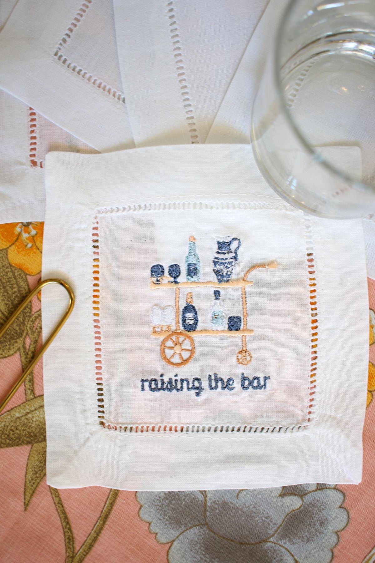 white hemstitch cocktail napkin with custom embroidery including a blue and gold bar cart and embroidered words below reading "raising the bar" in navy blue
