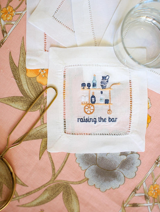 white hemstitch cocktail napkin with custom embroidery including a blue and gold bar cart and embroidered words below reading "raising the bar" in navy blue