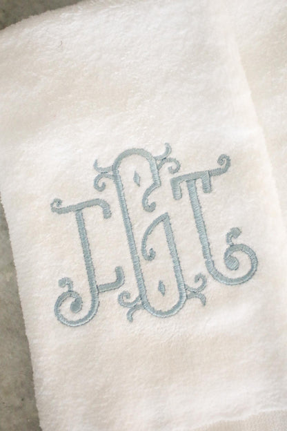 Detailed view of ornate triple-letter monogram embroidery in blue thread on white fingertip towel, custom personalized bathroom linen.