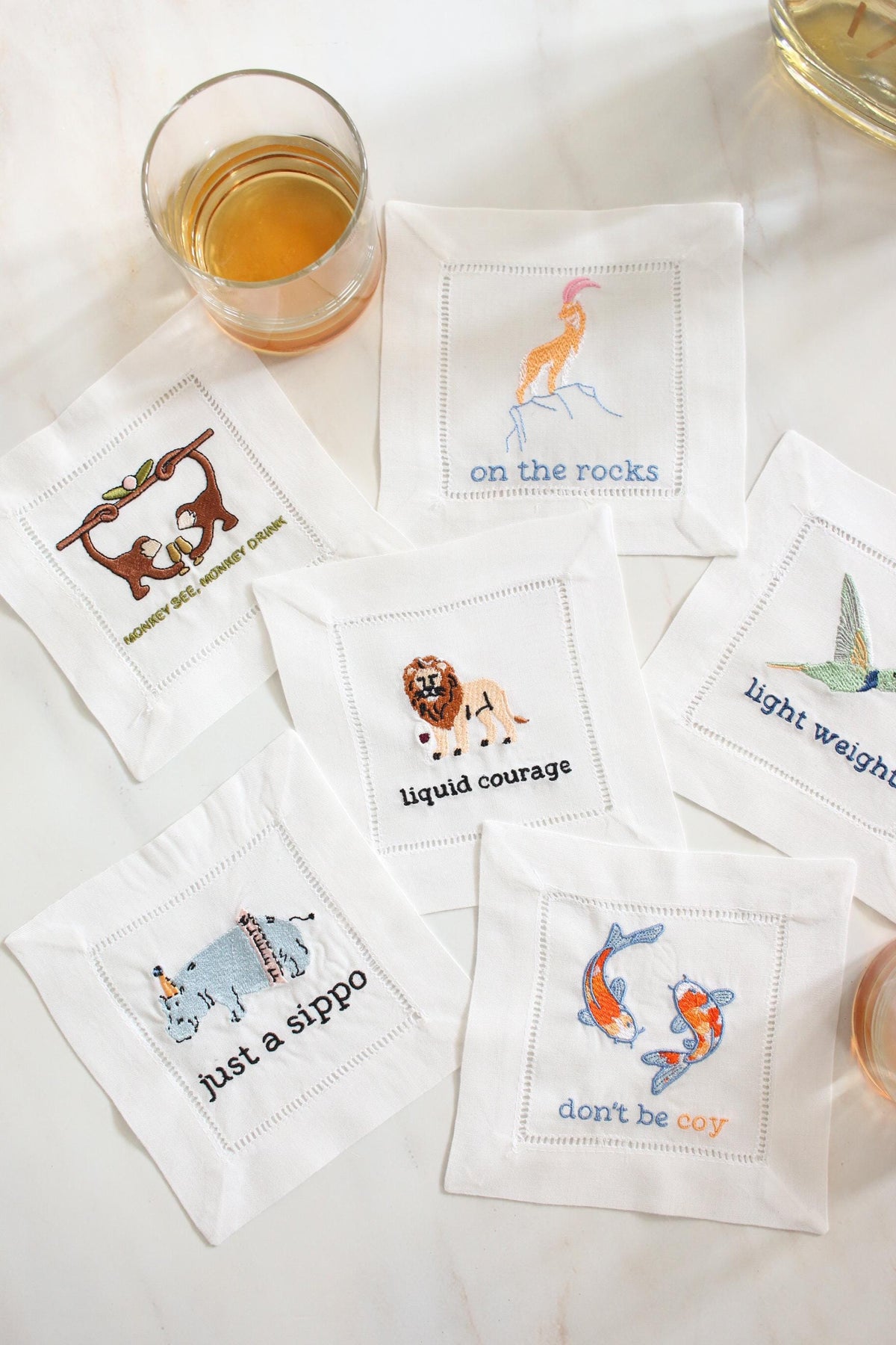 Set of six embroidered linen cocktail napkins displayed on a marble surface with funny animal cocktail designs