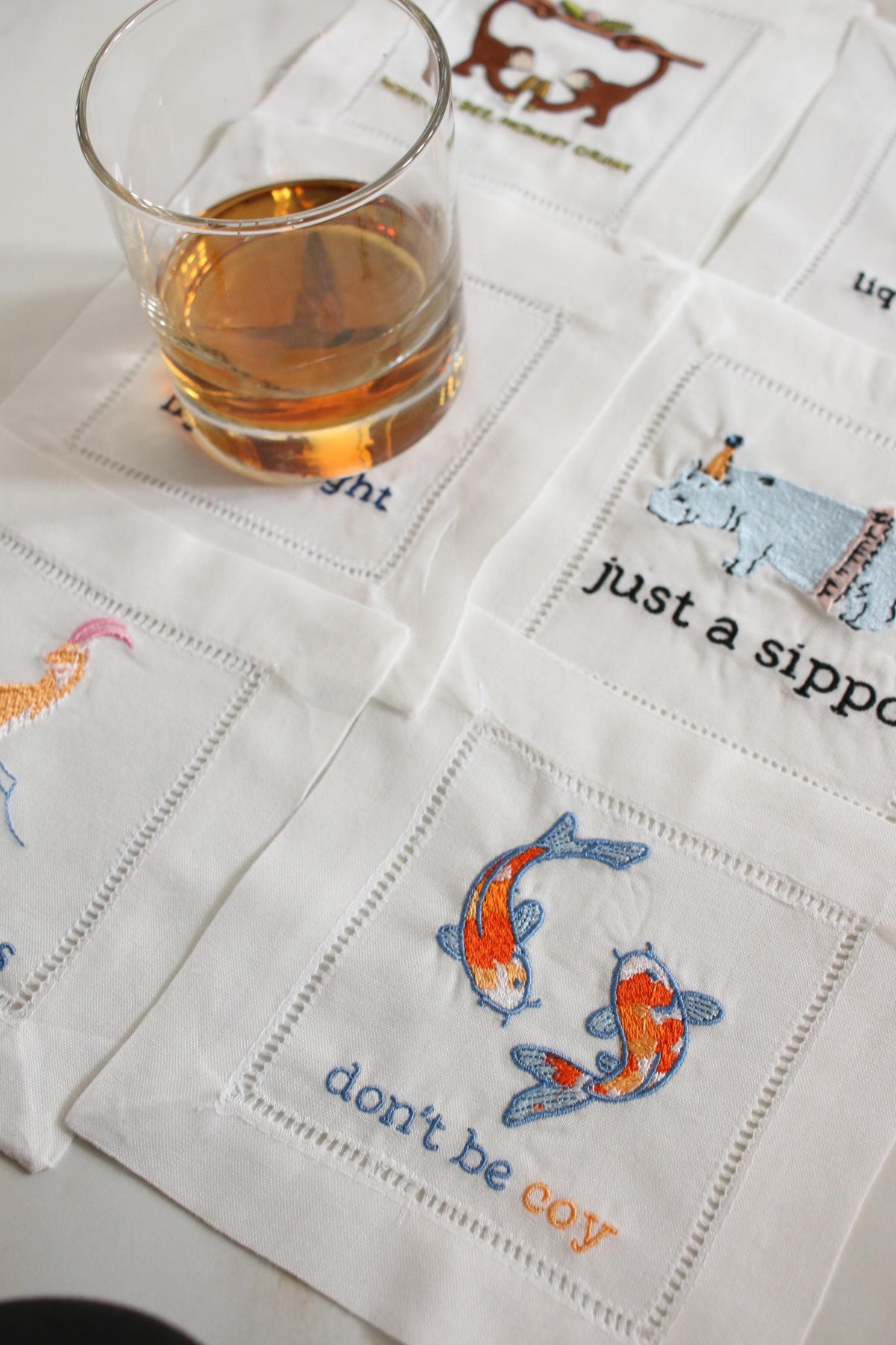 Funny embroidered linen cocktail napkins displayed with a whiskey glass on a marble bar